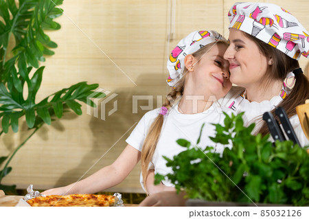 While eating home-made pizza, a five-year-old daughter hugs her mother warmly, thus reciprocating her mother's love for her child. 85032126