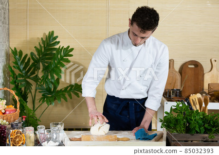 A young handsome chef kneads the dough in the kitchen with one hand on the stool that lies on the kitchen counter. A young handsome chef kneads the dough in the kitchen with one hand on the stool that lies on the kitchen counter. 85032393