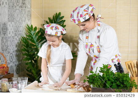 Mom and daughter roll out pizza dough with a wooden rolling pin on one board in the kitchen. Mother and baby do the dough together in the kitchen. Mom and daughter roll out pizza dough with a wooden rolling pin on one board in the kitchen. Mother and baby do the dough together in the kitchen. 85032429