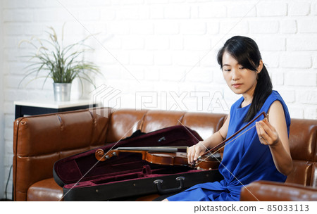 Young asian violinist in blue dress  takes care of the instrument after  practice session in living room. Rubbing rosin on the horse hair of the violin bow. 85033113