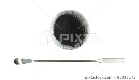 Carbon Charcoal Powder in Chemical Watch Glass placed next to the stainless spatula. Close up chemical ingredient on white laboratory table. Top View 85033172