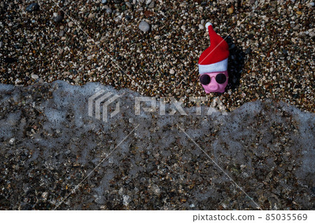Piggy bank in a hat of santa claus and sunglasses on a pebble beach by the sea. Piggy bank in a hat of santa claus and sunglasses on a pebble beach by the sea. 85035569