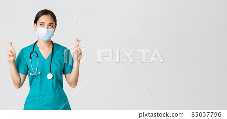 Covid-19, coronavirus disease, healthcare workers concept. Hopeful serious-looking asian doctor, female physician in medical mask and scrubs cross fingers, looking upper left corner Covid-19, coronavirus disease, healthcare workers concept. Hopeful serious-looking asian doctor, female physician in medical mask and scrubs cross fingers, looking upper left corner 85037796