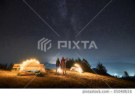 Boy standing near two bright tents, holding his girlfriend's hand and pointing her to scenery in front of him. Night camping on mountain meadow under starry sky. Concept of hiking, night camping. Boy standing near two bright tents, holding his girlfriend's hand and pointing her to scenery in front of him. Night camping on mountain meadow under starry sky. Concept of hiking, night camping. 85040658