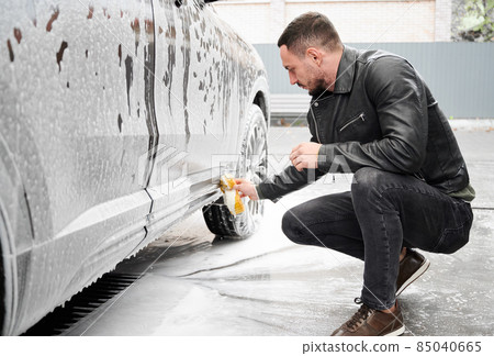 Young man washing car on carwash station outdoor. Handsome driver cleaning automobile, using sponge and soap. 85040665