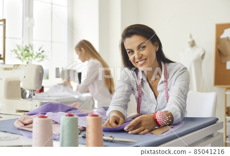 Portrait of woman dressmaker, fashion designer, tailor or seamstress at work in studio. Smiling woman looking at camera while working with fabric while sitting at her workplace in bright sewing studio Portrait of woman dressmaker, fashion designer, tailor or seamstress at work in studio. Smiling woman looking at camera while working with fabric while sitting at her workplace in bright sewing studio 85041216