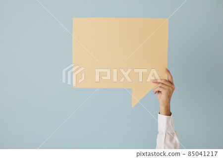 Unknown man raises his hand up holding speech bubble with copy space on light blue background. Close up of beige paper speech bubble layout for promotion, discount, advertisement or offer. 85041217