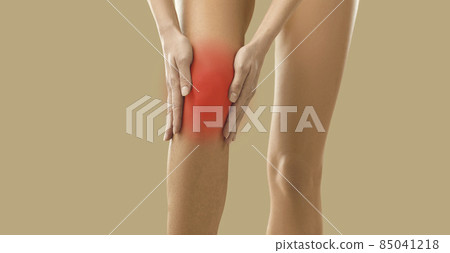 Close up on beige background woman holding sore knee where focus of pain is marked in red. Unknown young woman is experiencing joint pain, arthritis or tendon problems. Health concept. Medical banner. 85041218