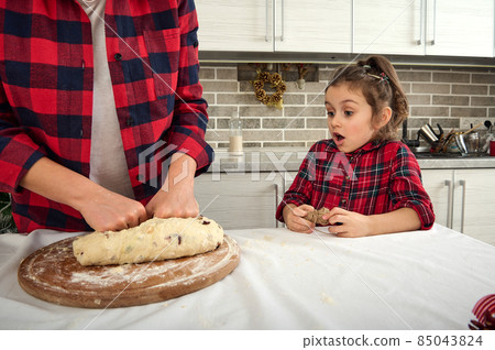 Close-up of a curious adorable little girl in checkered dress looking at her mother's hands kneading dough for Christmas cookies and bread 85043824