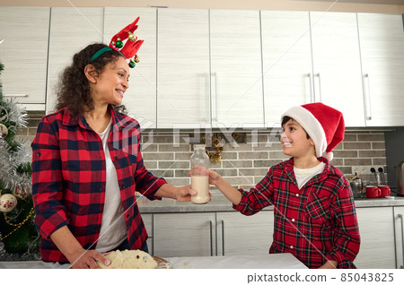 Handsome preadolescent boy in Santa hat bringing a bottle with milk to his loving mother cooking dough for Christmas cookies in the home kitchen. Son helping his mom in baking 85043825