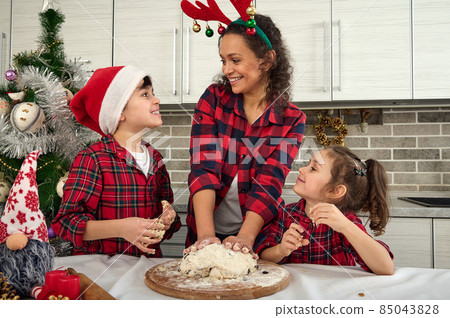 Happy mother with antler hoop on head prepares, kneads dough with raisins and nuts on a wooden board, smiles talking with her adorable kids, enjoys spending time together during Christmas holidays 85043828