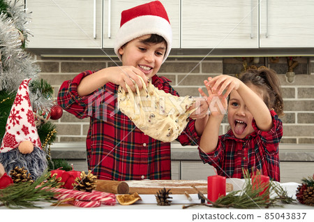 Beautiful children cooking together in the home kitchen during Christmas holidays. Adorable boy in Santa hat and his cute little sister preparing and kneading dough for bread 85043837