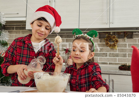 Cheerful brother and sister, adorable Caucasian children spend time together at home kitchen. Cute boy pours milk and baby girl kneads dough for Christmas cookies. Perfect happy family relationships 85043870