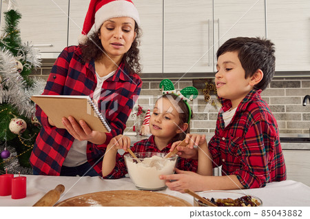 Happy cheerful family dressed in checkered clothes, beautiful young loving mom in Santa hat and two adorable children, boy and girl preparing together Christmas bread and cookies at home kitchen 85043882