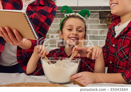 Close-up portrait of a happy cheerful sweet Caucasian baby girl smiles toothy smile looking at camera sitting at a kitchen table between her brother and mom while cooking together Christmas pastries Close-up portrait of a happy cheerful sweet Caucasian baby girl smiles toothy smile looking at camera sitting at a kitchen table between her brother and mom while cooking together Christmas pastries 85043884