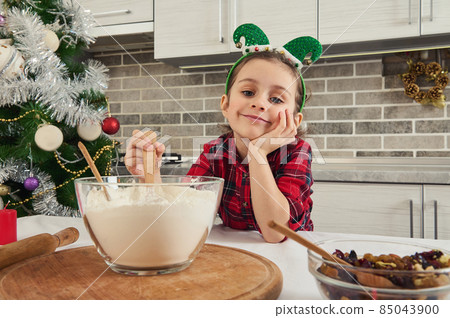 Adorable child, beautiful baby girl smiles, dreamily looking at the camera, mixing dry ingredients in a glass bowl. Cute little chef prepares cookies and sweets for christmas traditional events Adorable child, beautiful baby girl smiles, dreamily looking at the camera, mixing dry ingredients in a glass bowl. Cute little chef prepares cookies and sweets for christmas traditional events 85043900