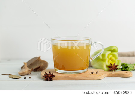 Meat broth with spices and vegetables in a mug, on a wooden board. Close-up. On a gray background, horizontal 85050926