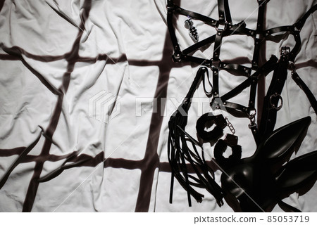 set of black leather sex toys for BDSM on the background of a sheet in bed. Whip, handcuffs, belt belt and mask 85053719