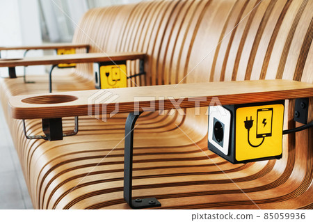 A designer wooden bench with sockets and a yellow sign for charging smartphones in the waiting room. A designer wooden bench with sockets and a yellow sign for charging smartphones in the waiting room. 85059936