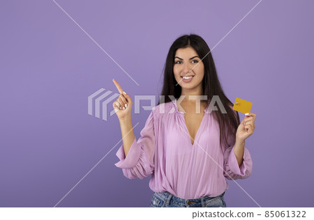 Happy armenian lady with credit card pointing aside at free space and smiling at camera over violet studio background Happy armenian lady with credit card pointing aside at free space and smiling at camera over violet studio background 85061322
