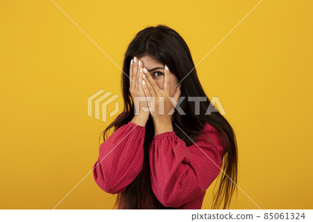 Frightened young armenian lady trying to hide, covering face with hands and peeking with one eye, yellow background Frightened young armenian lady trying to hide, covering face with hands and peeking with one eye, yellow background 85061324