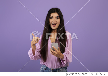 Yes, great news. Surprised armenian woman celebrating win, using smartphone and pointing at gadget, purple background Yes, great news. Surprised armenian woman celebrating win, using smartphone and pointing at gadget, purple background 85061507