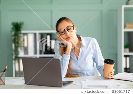 Young businessman tired of computer work sleeping at workplace, holding coffee cup in hand Young businessman tired of computer work sleeping at workplace, holding coffee cup in hand 85061717