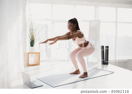 Full length portrait of happy young black woman doing squats in front of laptop at home, free space 85062118