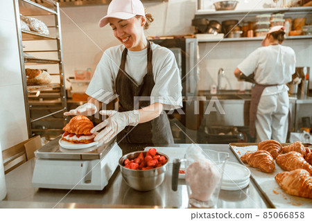 Smiling woman with croissant with strawberry and cream on digital scales in bakery 85066858