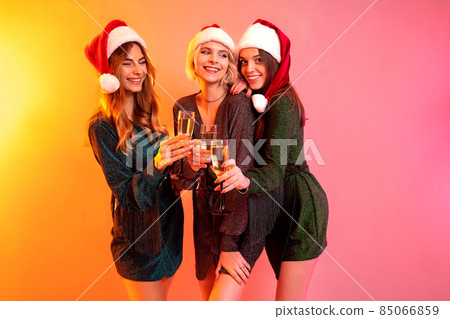 Three happy girls in Santa Claus hats holds glasses of champagne having fun. New Year party 85066859