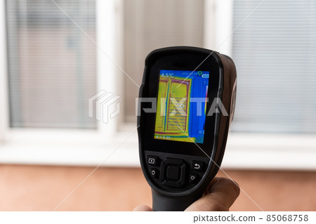 A male hand holds a thermal imager at the window of a house. Search for heat loss in private houses. 85068758
