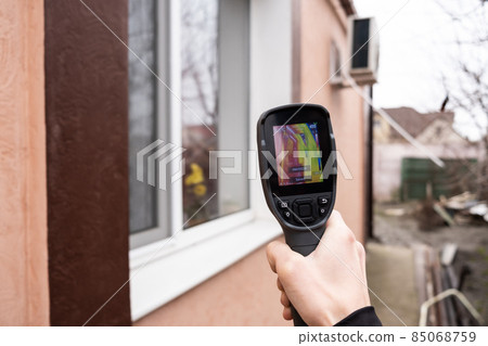 A male hand holds a thermal imager at the window of a house. Search for heat loss in private houses. A male hand holds a thermal imager at the window of a house. Search for heat loss in private houses. 85068759