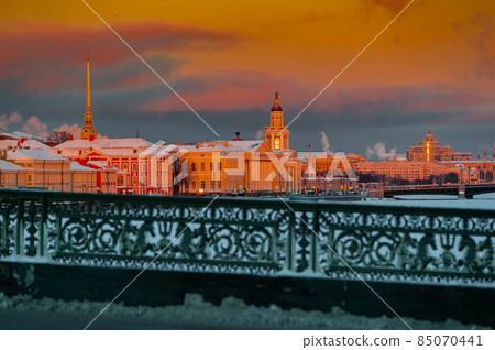 Winter panoramic view of St. Petersburg at sunset, golden spire of Peter and Paul fortress, Kunstkamera tower, embankment with bridge on background, steam over frozen Neva river, sky of orange color Winter panoramic view of St. Petersburg at sunset, golden spire of Peter and Paul fortress, Kunstkamera tower, embankment with bridge on background, steam over frozen Neva river, sky of orange color 85070441