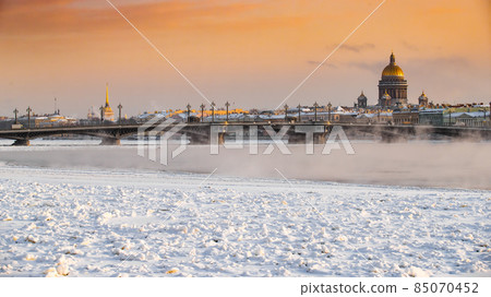 Winter panoramic view of St. Petersburg at sunset, Isaac cathedral and Blagoveshenskiy bridge on background, steam over frozen Neva river, sky of orange color 85070452