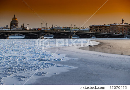 Winter panoramic view of St. Petersburg at sunset, Isaac cathedral and Blagoveshenskiy bridge on background, steam over frozen Neva river, sky of orange color Winter panoramic view of St. Petersburg at sunset, Isaac cathedral and Blagoveshenskiy bridge on background, steam over frozen Neva river, sky of orange color 85070453