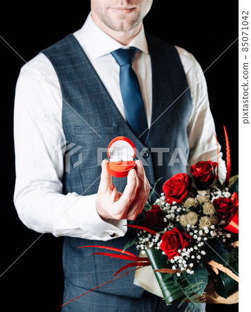 Marriage ring. Young happy man holding red gift box with diamond engagement ring, red roses bouquet. Love propose, couple relationship with copy space. 85071042