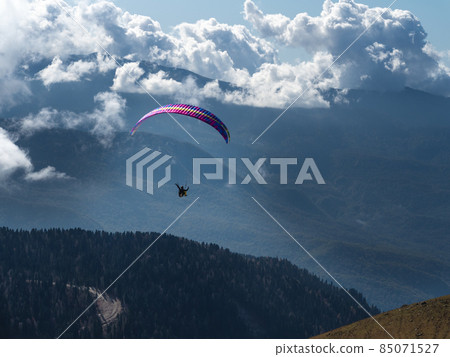 A man on a paraglider flies through the clouds over the forest and mountains. 85071527