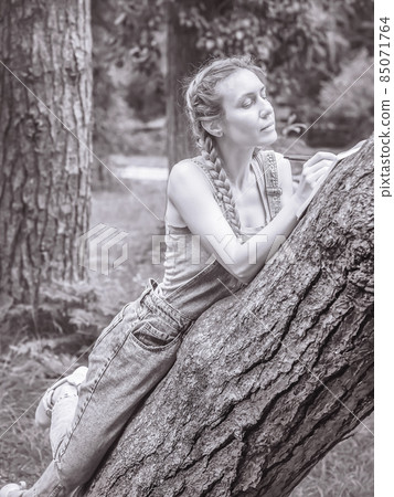 A woman makes notes in a notebook leaning on a tree trunk in the park. Black and white photo A woman makes notes in a notebook leaning on a tree trunk in the park. Black and white photo 85071764