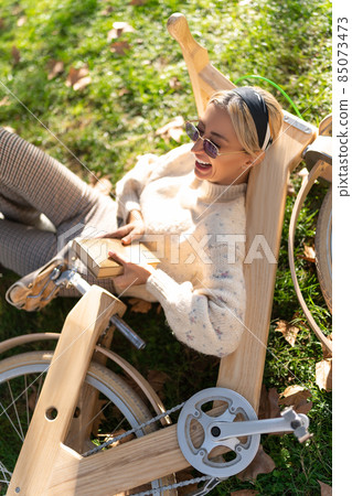 From above of female in sunglasses laughing with opened mouth while lying on grass and leaning on timber bicycle in park 85073473