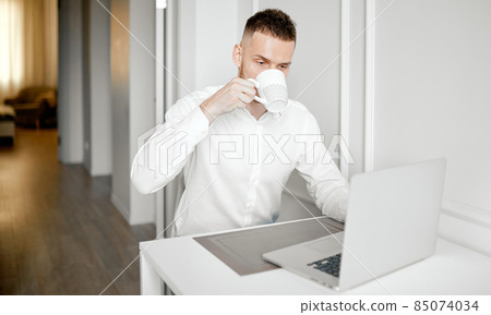 A man drinks coffee working at a laptop in his kitchen 85074034