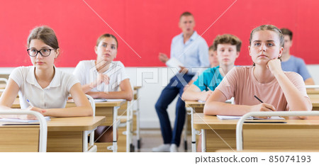 Focused teenage students studying in classroom with teacher, writing lectures in workbooks Focused teenage students studying in classroom with teacher, writing lectures in workbooks 85074193