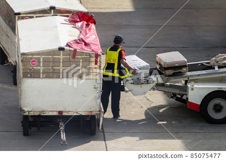 asian man loader lifting up the luggage to conveyor belt of the trailer to loading passenger baggage to the airplane on the runway at the airport. travel, transportation and logistics concept. 85075477