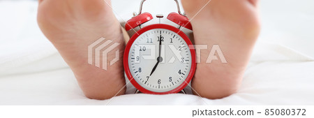 Red alarm clock standing in bed between female heels closeup Red alarm clock standing in bed between female heels closeup 85080372