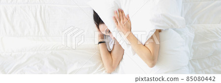 Young woman lying in white bed and covering her ears with pillow top view 85080442