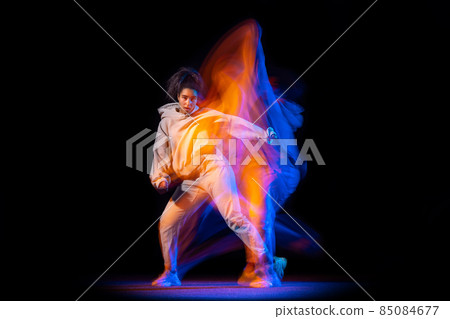 Sportive young girl in white costume dancing hip-hop dance isolated on dark background at dance hall in neon mixed light. Sportive young girl in white costume dancing hip-hop dance isolated on dark background at dance hall in neon mixed light. 85084677