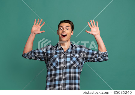 Half-length portrait of young shocked man, student standing with raising hands isolated on green studio background. 85085240