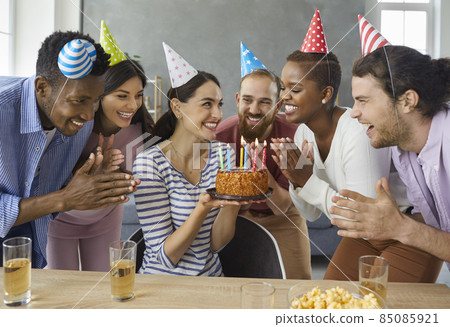 Group of cheerful young multiracial people in conical party hats applaud while the birthday girl makes a wish. Happy woman holding a birthday cake while celebrating at home with her best friends. Group of cheerful young multiracial people in conical party hats applaud while the birthday girl makes a wish. Happy woman holding a birthday cake while celebrating at home with her best friends. 85085921