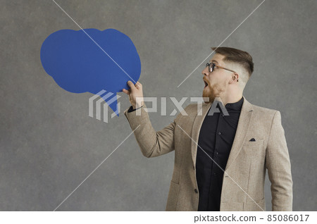 Young adult businessman looking at blank chat message speech bubble with surprised emotion standing over grey background. Positively shocked guy showing wow expression on face with mouth opened Young adult businessman looking at blank chat message speech bubble with surprised emotion standing over grey background. Positively shocked guy showing wow expression on face with mouth opened 85086017
