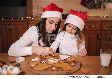 Mom in santas hat with her little doughter decorates gingerbread using glaze. Christmas and New Year traditions concept. Christmas bakery. Happy holidays 85086889