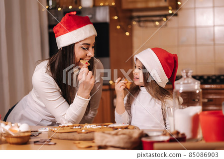Adorable little girl and her mom in santa hat eating homemade gingerbread and laughing. Happy little kid preparing for Christmas. Background of fairy lights bokeh Adorable little girl and her mom in santa hat eating homemade gingerbread and laughing. Happy little kid preparing for Christmas. Background of fairy lights bokeh 85086903